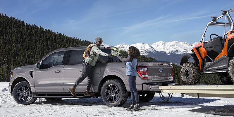 Ford pickup truck in snowy mountains with two people and an off-road vehicle on a trailer