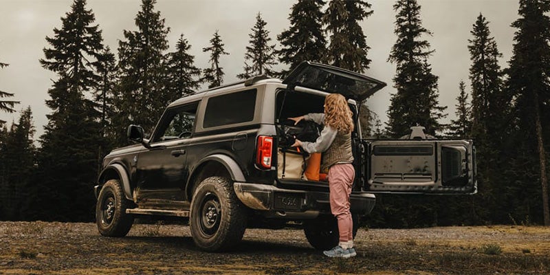 Woman unloading gear from the back of an SUV in a forest setting.