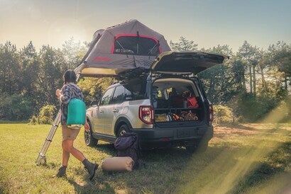 A Ford Bronco Sport parked on a grass field with camping gear in the trunk, a tent on top, and a woman walking by