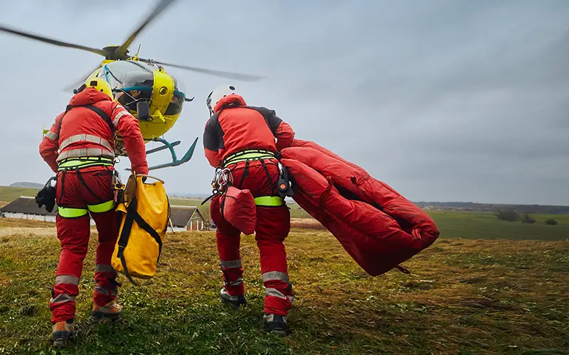 Two rescue workers approach a helicopter with emergency gear in an open field.